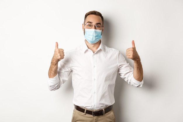 Covid-19, social distancing and quarantine concept. Satisfied male manager showing thumbs up, recommending to wear face mask, standing over white background