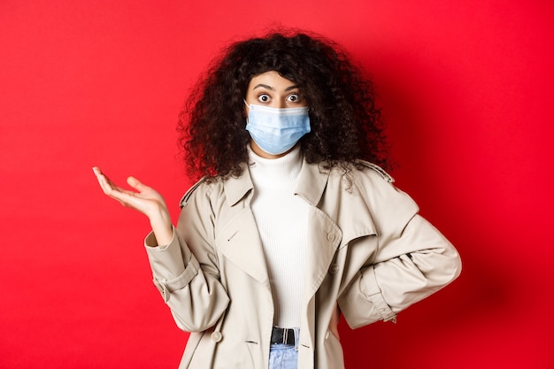 Covid-19, social distancing and quarantine concept. Shocked and confused european woman with curly hair, wearing medical mask from coronavirus, raise hand up puzzled, red background.