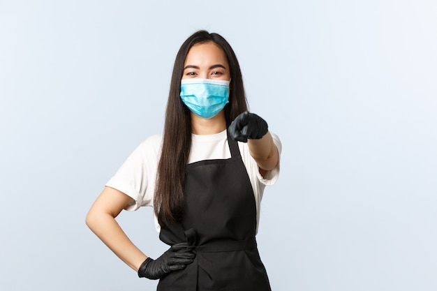 Covid-19, social distancing, small coffee shop business and preventing virus concept. Cheerful asian employee, restaurant barista in medical mask and gloves pointing at camera, invite consumers