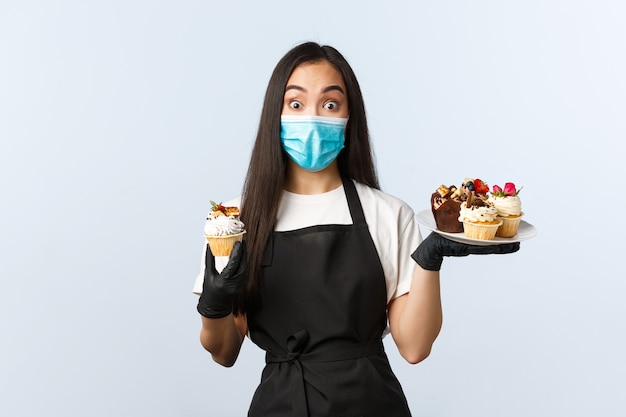 Covid-19, социальное дистанцирование, небольшая кофейня и концепция предотвращения вирусов. Взволнованная милая азиатская женщина-бариста рекомендует новое летнее меню десертов, держит в руках кексы и надевает медицинскую маску.