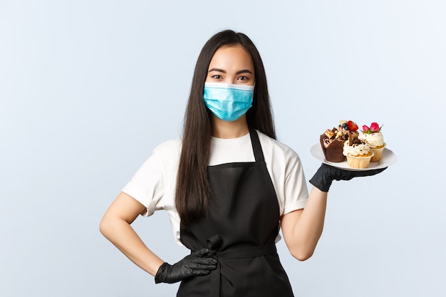 Covid-19, sociale afstand, kleine coffeeshopzaken en het voorkomen van virusconcept. leuke aziatische serveerster in café, werknemer met medisch masker en handschoenen die desserts serveren, bord met muffins vasthouden