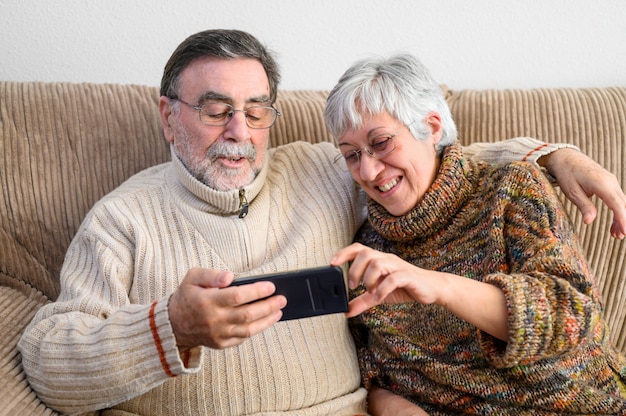 Covid-19は家にいます。幸せな引退した年配のカップル、携帯電話で家族のビデオ通話を行います。社会的距離、肯定的な表現。