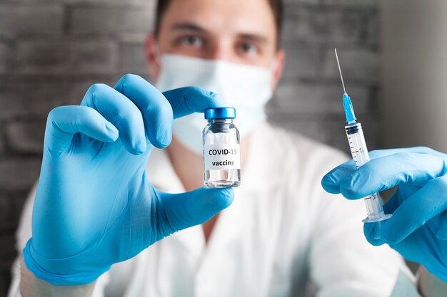 COVID-19 vaccine in researcher hands, male doctor in a white coat, in a medical mask, in blue gloves holds syringe and bottle with vaccine for coronavirus cure. Concept of corona virus treatment.