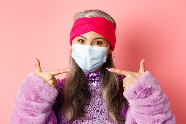 Photo covid-19, virus and fashion concept. fashionable asian senior woman in trendy outfit pointing fingers at medical mask, follow social distancing rules, pink background