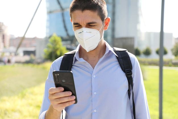 COVID-19 Young business man wearing KN95 protective mask using smartphone app in modern city street