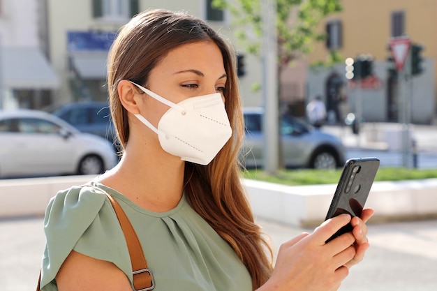 Foto covid-19 giovane donna che indossa la maschera ffp2 utilizzando uno smartphone in una strada cittadina