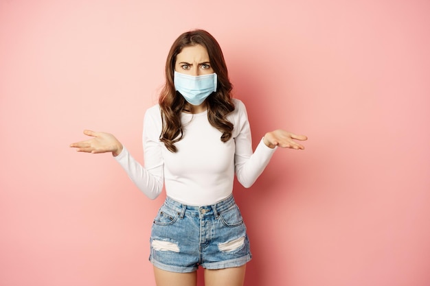 Covid confused young woman in medical face mask shrugging shoulders and looking clueless standing pu...