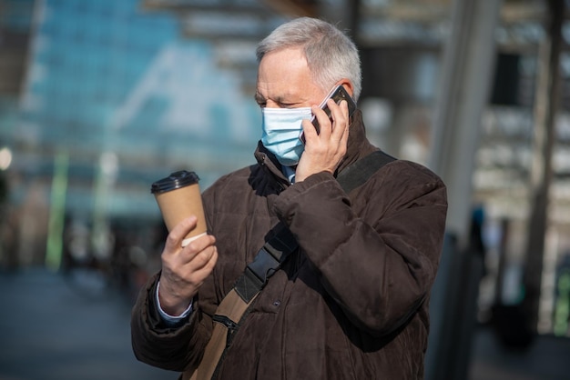 Covid coronavirus concept gemaskerde oudere man die buiten op zijn smartphone praat en een kopje koffie houdt