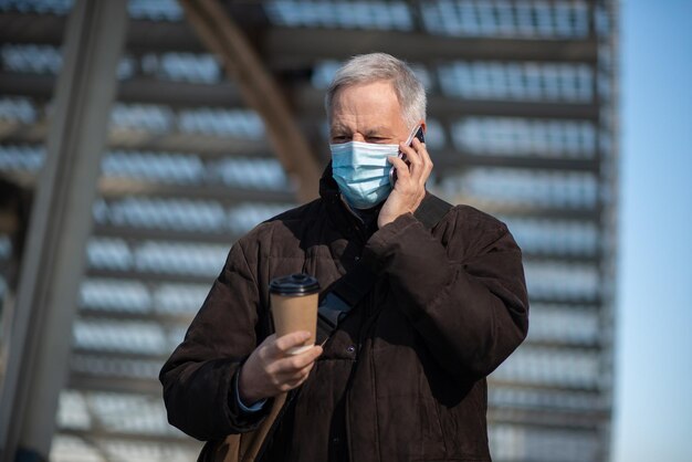 Covid coronavirus concept gemaskerde oudere man die buiten op zijn smartphone praat en een kopje koffie vasthoudt