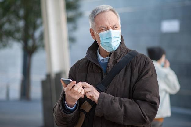 Covid coronavirus concept gemaskerde oudere man met behulp van zijn smartphone buiten