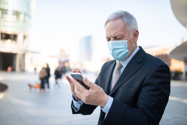 Covid coronavirus concept, masked elder businessman using his smartphone outdoor