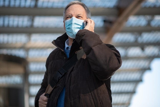 Covid coronavirus concept masked elder man talking on his smartphone outdoor