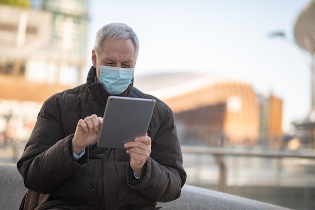 Covid coronavirus levensstijl gemaskerde oudere man die zijn tablet gebruikt terwijl hij buiten op een stadsplein zit