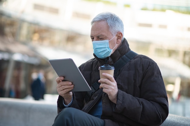 Covid coronavirus levensstijl gemaskerde oudere man die zijn tablet gebruikt terwijl hij buiten op een stadsplein zit