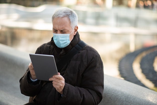 Covid coronavirus levensstijl, gemaskerde oudere zakenman met behulp van zijn tablet terwijl hij buiten op een stadsplein zit