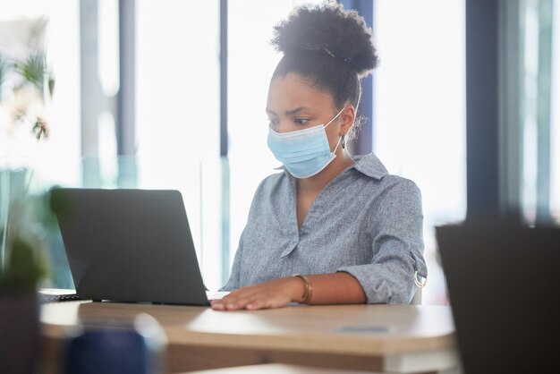 Covid face mask and a black woman working on a laptop in a modern office Sick businesswoman on the internet at work College student online exam and in campus library protected against the virus