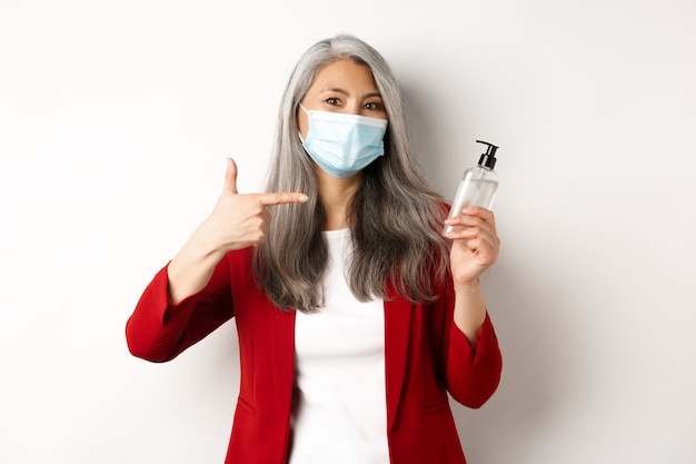 Covid, pandemia e concetto di business. manager femminile asiatico in maschera facciale, puntando il dito a portata di mano disinfettante, raccomandando antisettico, sfondo bianco