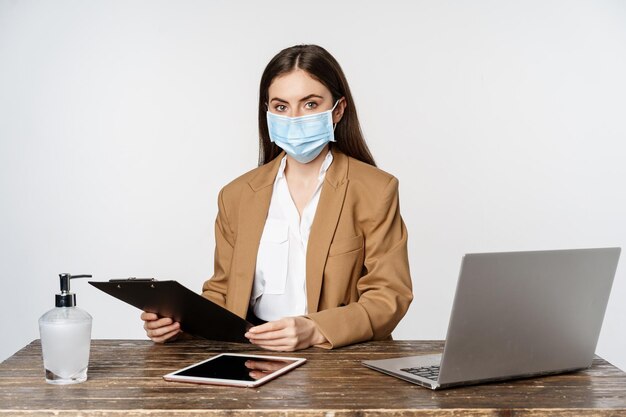 Covid, pandemic and office workplace concept. Portrait of corporate woman in medical face mask from coronavirus, working, standing over white background