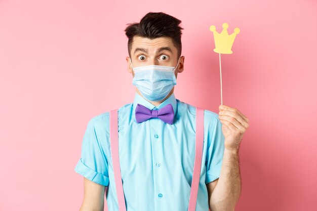 Foto covid, pandemia e concetto di quarantena. ragazzo carino e sciocco in maschera medica che sembra sorpreso, tenendo in mano una piccola corona per la festa, in piedi su sfondo rosa.