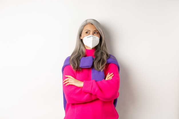 Covid, pandemic and social distancing concept. Stylish asian middle-aged woman with grey hair, wearing face mask and cross arms on chest confident, looking upper left corner, white background.