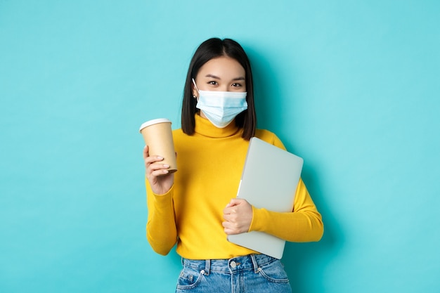 Covid, пандемия и концепция социального дистанцирования. Стильная азиатская женщина в медицинской маске, держа чашку кофе и ноутбук, идя на работу, стоя на синем фоне.