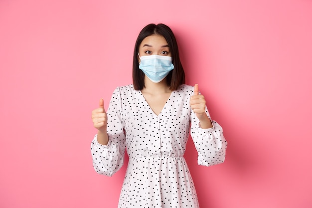 Covid quarantine and lifestyle concept excited asian woman in face mask showing thumbsup praising go...