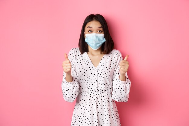 Covid quarantine and lifestyle concept excited asian woman in face mask showing thumbsup praising go...