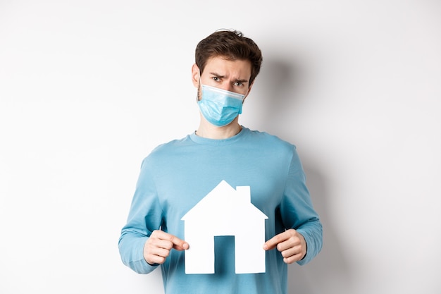 Covid and real estate concept. Sad and doubtful young man in medical mask feeling reluctant, showing paper home cutout, standing over white background.