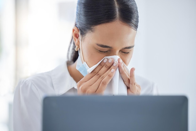 Covid sick and blowing nose with a business woman sneezing into a tissue while suffering with a cold flu or allergies in the office Hands nasal and hayfever with a young female employee at work