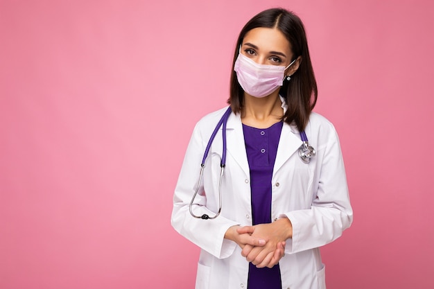 Covid19, coronavirus and doctors concept. Photo of professional confident young european doctor in medical mask and white coat, stethoscope over neck isolated over pink wall. Empty space