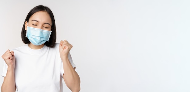 Covid19 healthcare and medical concept Enthusiastic asian woman in medical face mask dancing and celebrating winning achieve goal standing over white background