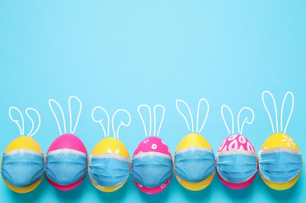 Photo covid19 pandemic colorful easter eggs with cute bunny ears in protective masks and space for text on light blue background flat lay