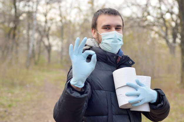 Концепция Ковидиот. Молодой человек в защитной маске держит много рулонов туалетной бумаги и показывает хорошо жест на открытом воздухе в весеннем лесу