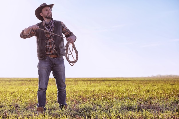 Cowboy in a hat and with a lasso standing in a field at sunset