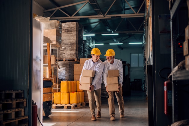 Coworkers carrying boxes with helmets on heads. Storage interior.