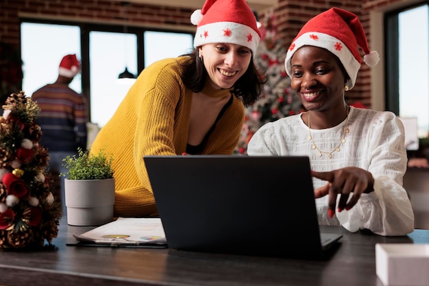 お祝いの会社のオフィスでチームワークを行い、ラップトップで作業して、クリスマスの飾りやライトで満たされたスペースでプロジェクトを計画する同僚.クリスマス ツリーと装飾のオフィスの仕事の人々。