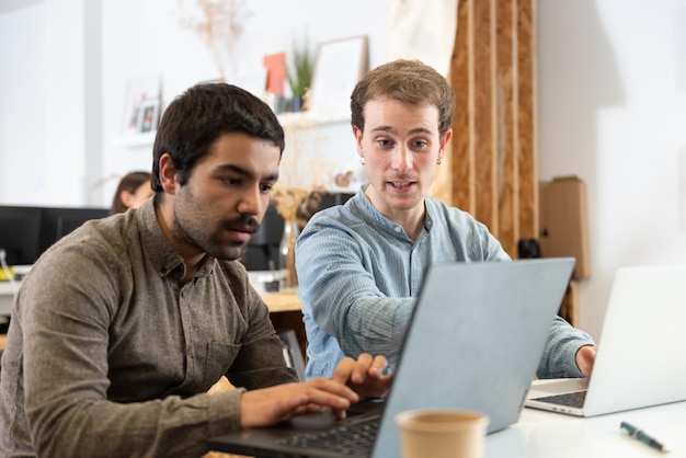 Coworkers helping each other while working on their laptops in the office.