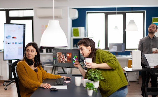 Photo coworkers talking about movie project looking at film footage working in creative start up agency office with two monitors