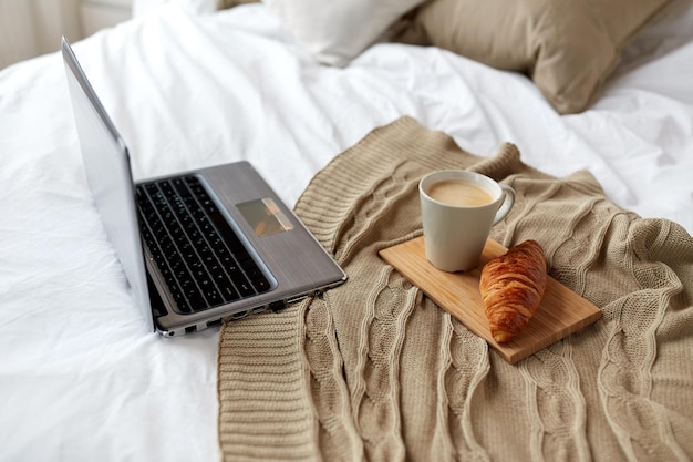 coziness, technology, interior and winter concept - cozy bedroom with laptop computer, coffee cup and croissant on bed at home