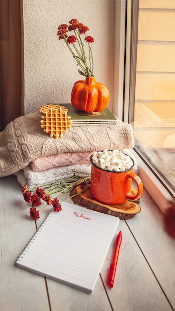 Фото Уютный осенний натюрморт на подоконнике с блокнотом для заметок: теплые шерстяные кофточки, тыквы, кленовые листья и чашка какао с зефиром и вафлями.