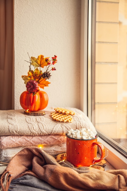 Photo cozy autumn still life on the windowsill: warm wool sweaters, scarf, pumpkins, maple leaves and a cup of cocoa with marshmallows and waffles.