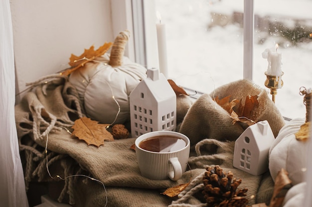 Cozy Autumn Warm cup of tea stylish pumpkin pillows fall leaves candle lights and cute buildings decoration on brown scarf on windowsill Autumn hygge fall home decor