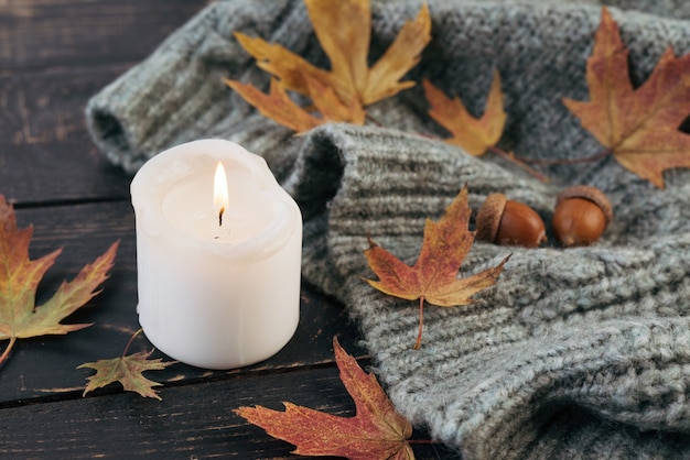 Cozy and autumnal atmosphere. A candle is burning against the background of a knitted blanket with fallen autumn leaves on a dark wooden table