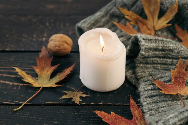 Cozy and autumnal atmosphere. A candle is burning against the background of a knitted blanket with fallen autumn leaves on a dark wooden table