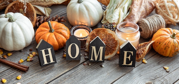 Cozy autumnal composition with decorative word home, candles, pumpkins, corn on a wooden surface in a rustic style.