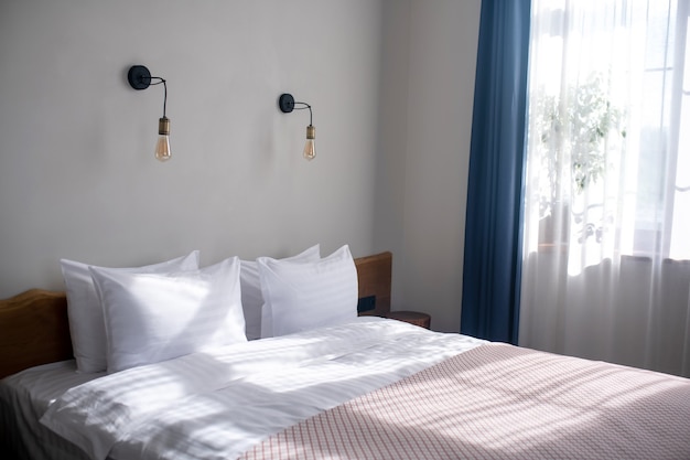 Уютная спальня. Большая удобная кровать с белыми подушками и покрывалом в светлой спальне, освещенной солнечным светом, и окном.
