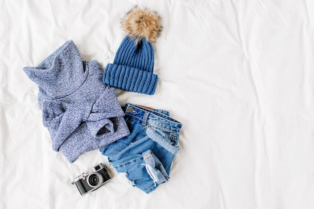 Cozy blue sweater,  jeans,  hat and photo camera on bed on white sheet. Women's stylish autumn or winter outfit. Trendy  clothes collage. Flat lay, top view.