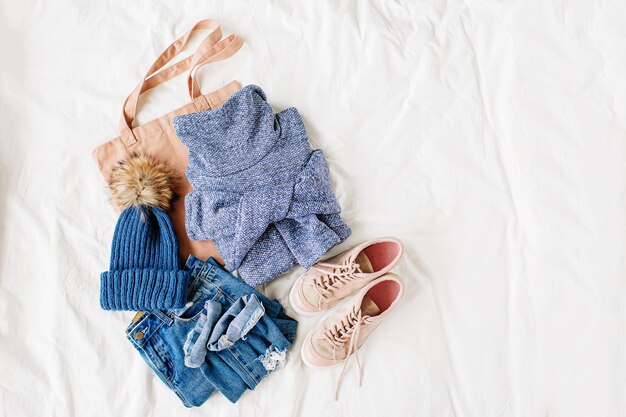 Cozy blue sweater,  jeans, sneakers, and hat on bed on white sheet. Women's stylish autumn or winter outfit. Trendy  clothes collage. Flat lay, top view.