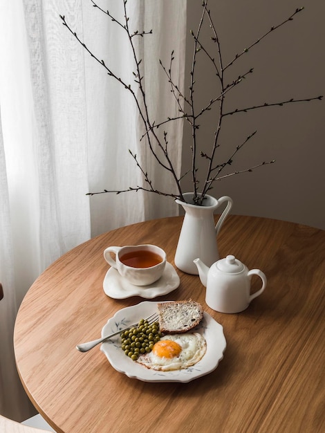 Уютный завтрак, бранч, чай, жареное яйцо с консервированным горохом, хлебом и маслом на круглом деревянном столе.