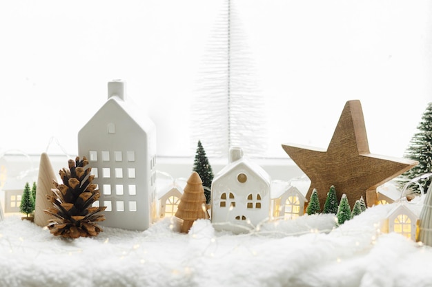 Cozy christmas miniature village Stylish cute little ceramic houses and wooden trees on soft snow blanket with glowing lights in room Atmospheric winter village still life Merry Christmas